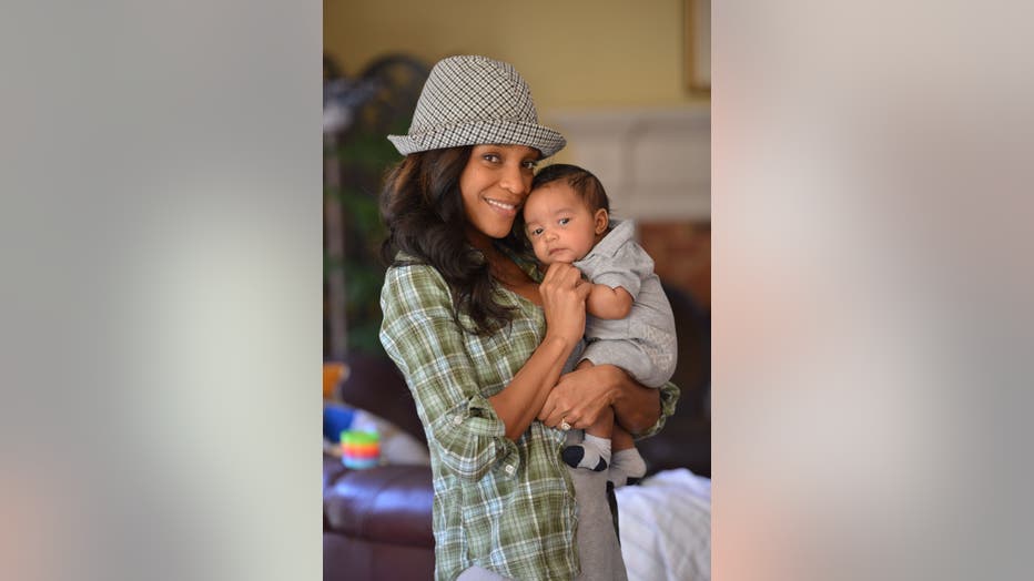 African American woman wearing a hat and a plaid shirt hugs her infant son as she smiles.