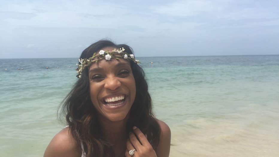 Woman stands on the beach, with her fingers in her hair. She's wearing a crown of flowers and is laughing.