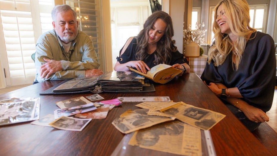 Bearded man and his two adult daughters sit at a table and look through newspaper clippings from 1972 detailing his kidney transplant.