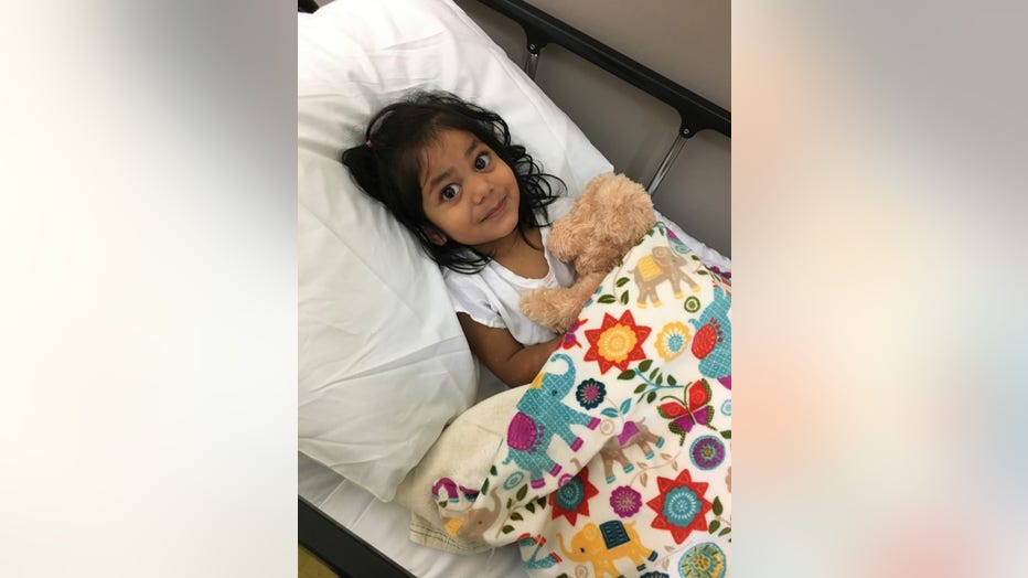 A little girl with curly dark hair smiles up from a hospital bed. She is holding a stuffed animal.