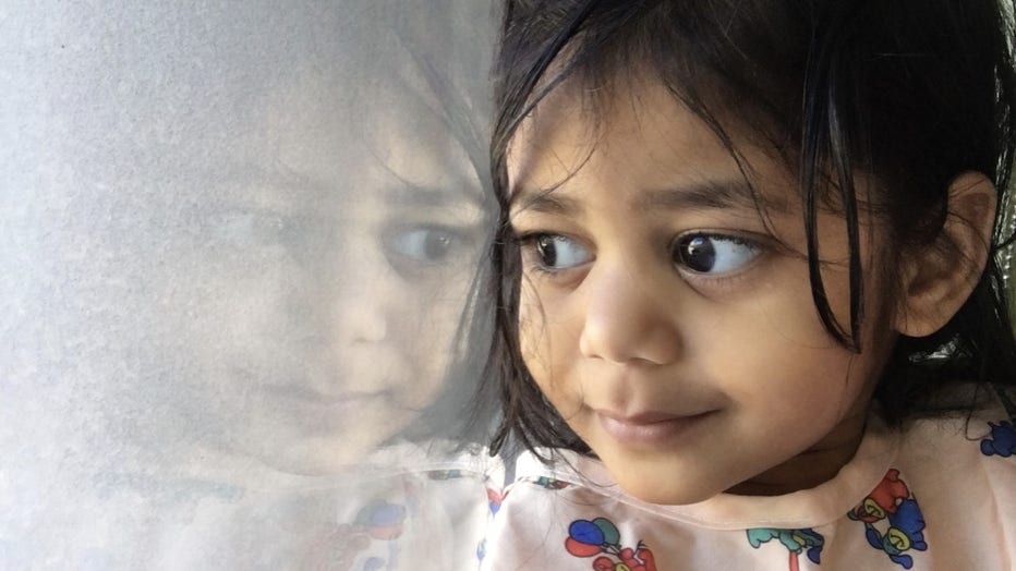 A little girl with dark hair and large eyes pushes her face against a hospital window. She's wearing a hospital gown and is smiling.