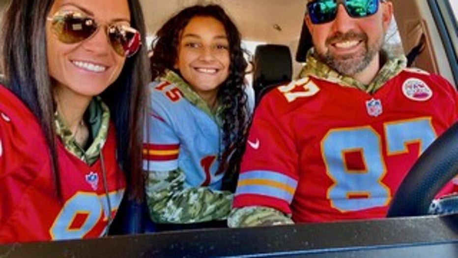 Man and woman wearing football jerseys sit in their vehicle with a girl, the woman's daughter, sitting between them.