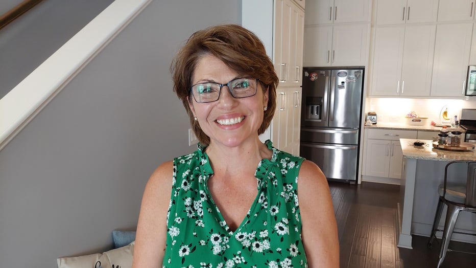 Woman with short hair and a flowered dress smiles while standing in her living room.