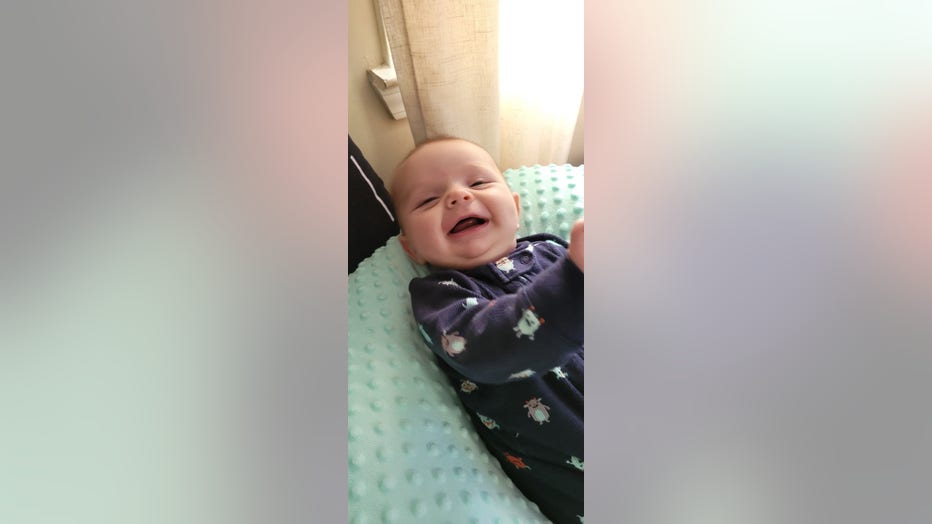 A baby boy laughs at the camera as his mom takes a photo. He's lying with his back on a blanket.