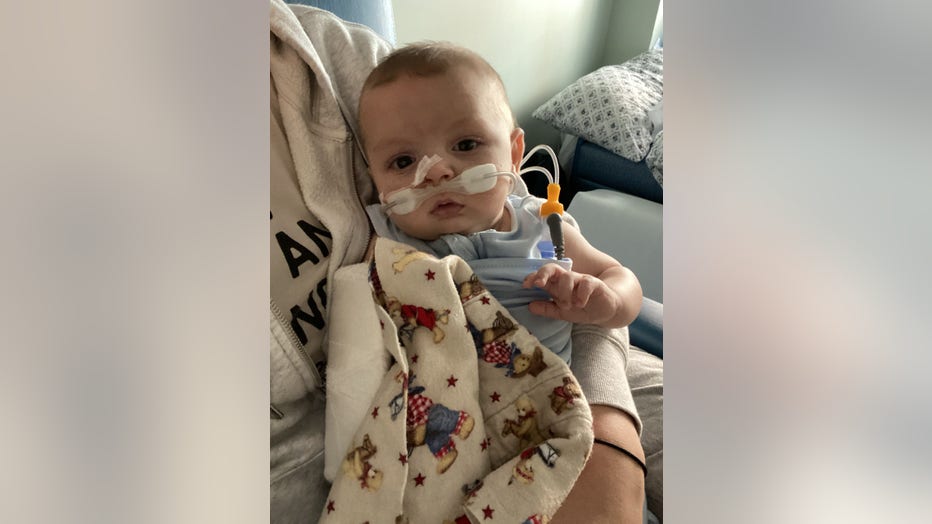 A baby with RSV sits in his mother's lap in the hospital. He has a feeding tube and an oxygen tube taped to his nose. 