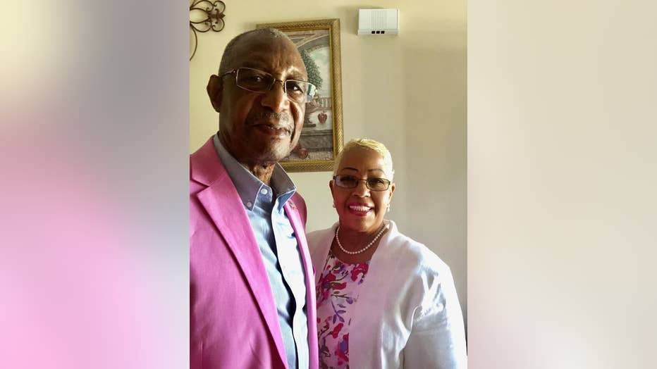 Woman stands next to her husband. She is smiling and wearing a white jacket and pearls.