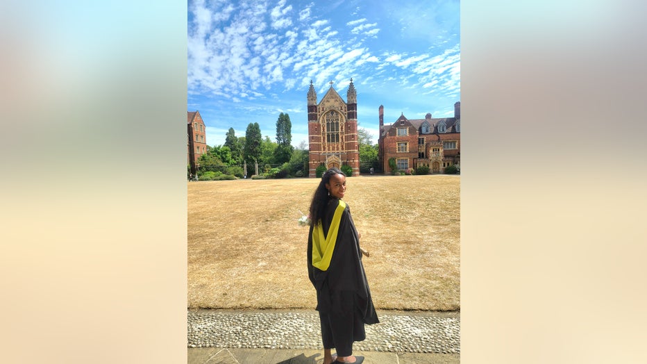 Young African American woman wearing a graduation gown stands in front of buildings at Cambridge University.