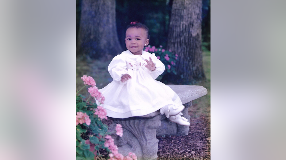 A small girl, about 18 months, poses while sitting on a bench. Her hair is short and she is wearing a white dress. 