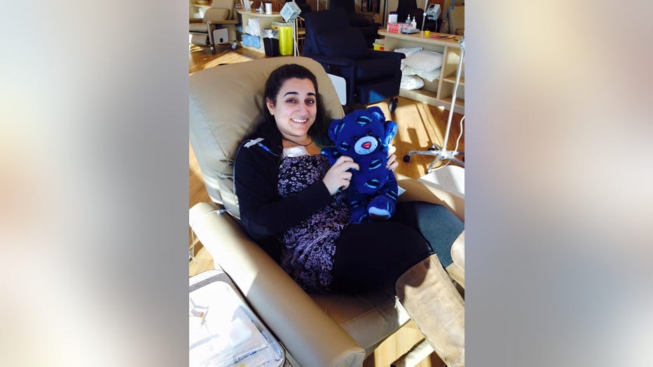 Woman with dark hair hugs a teddy bear while sitting in a treatment chair during a chemotherapy infusion.