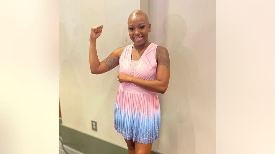 April Addison, with her head bald from chemotherapy, holds up her right arm and shows her muscles as she smiles at the camera.