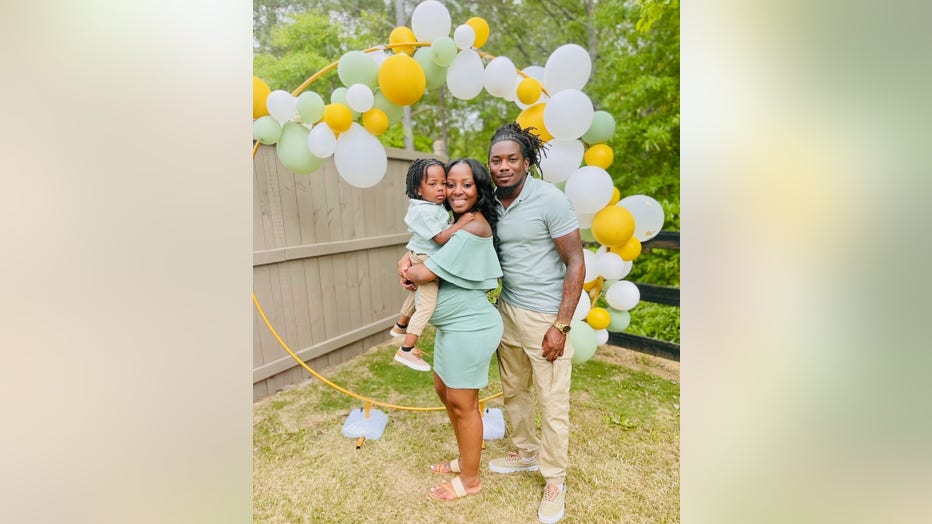 A couple stands in front of a circle of balloons at their baby shower. They are holding their toddler and smiling.