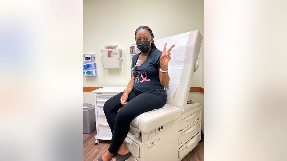 April Addison makes a peace sign as she sits in an exam room. On her chest, you can see she has a chemotherapy port. She's wearing a mask and looks confident.