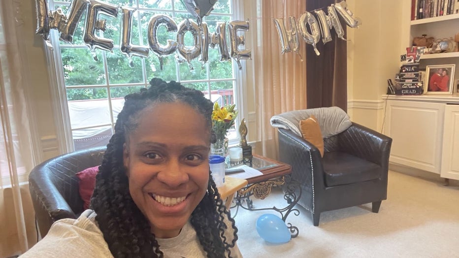 Woman poses in her living room in from of a balloon sign that reads, "Welcome Home."