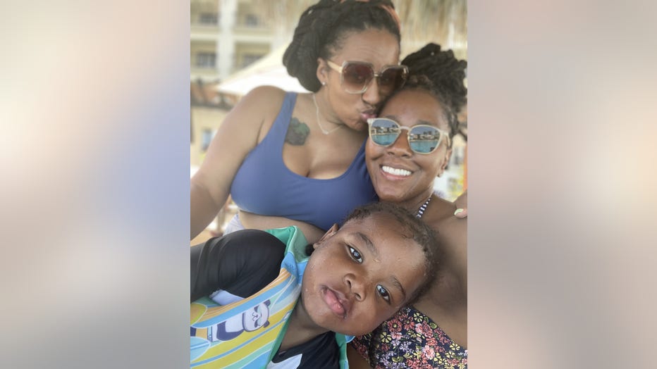 Two Black women wearing sunglasses and bathing suits pose poolside with their young son