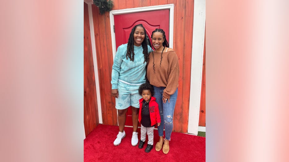 A Black couple in their thirties, both women, stand with their young son between them. They are smiling.