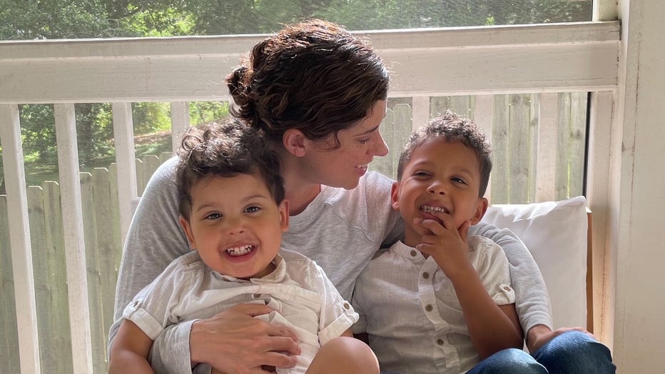 A mother sits between her two young children on a sunny porch. She is hugging them , with her face in profile as she looks at one of them.