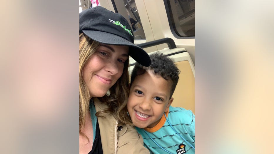 A woman wearing a shoulder length wig and baseball cap smiles as she takes a photo with her young son