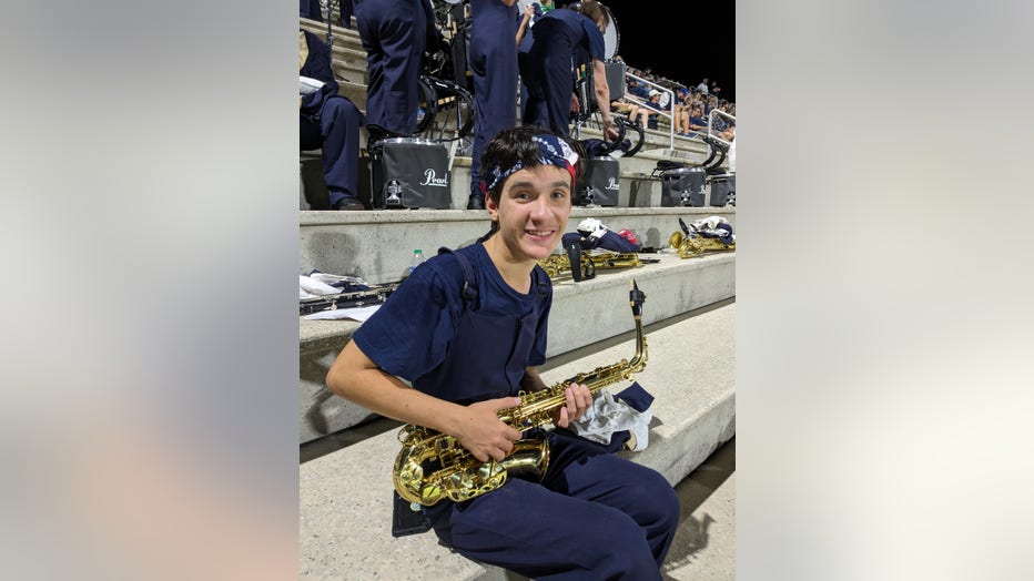 Dark haired teenage boy sits in the bleachers holding his saxophone at a football game. Hes smiling at the camera.