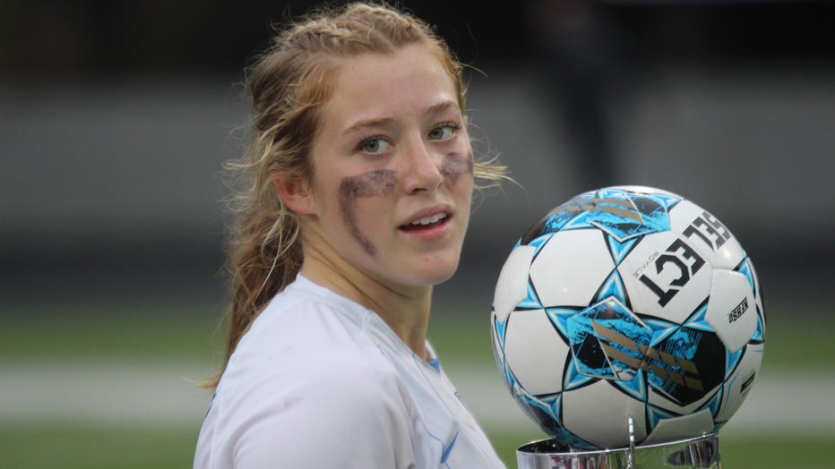 Teenage girl in soccer jersey, with her face painted, holds a soccer ball as she looks back at the camera.