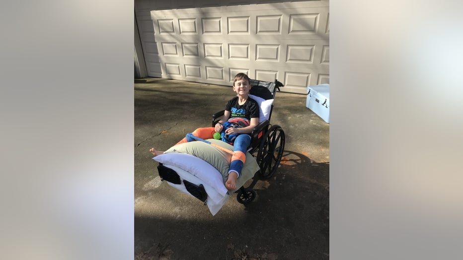 Boy sits in wheelchair wearing a V shaped lower body cast that holds his legs apart after undergoing major hip surgery. Hes smiling as his back and legs are propped up on pillows.