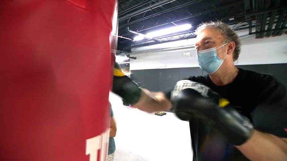 A 67-year-old man with curly hair punches a boxing bag during a non-contact boxing class for patients with Parkinsons disease. He is wearing a face mask because the photo was taken in June of 2022, just as pandemic restrictions began to ease.