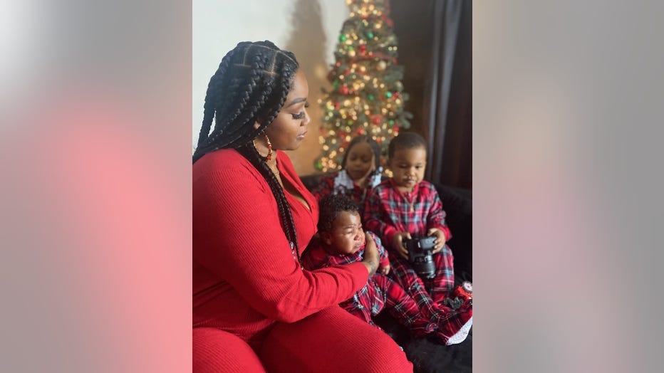 A mother holds her baby son in her lap. In the background, you can see her two other children and a Christmas tree.