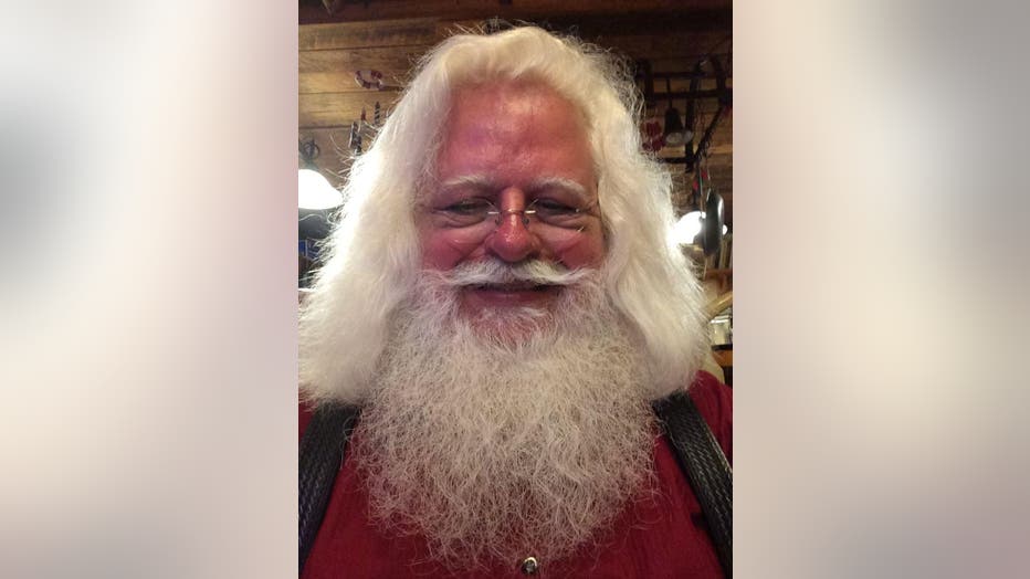 A tight shot of Jonathan Byrd's face in his Santa Clause costume. He has long white hair and a fluffy beard and is wearing small Santa-like glasses and a red shirt.