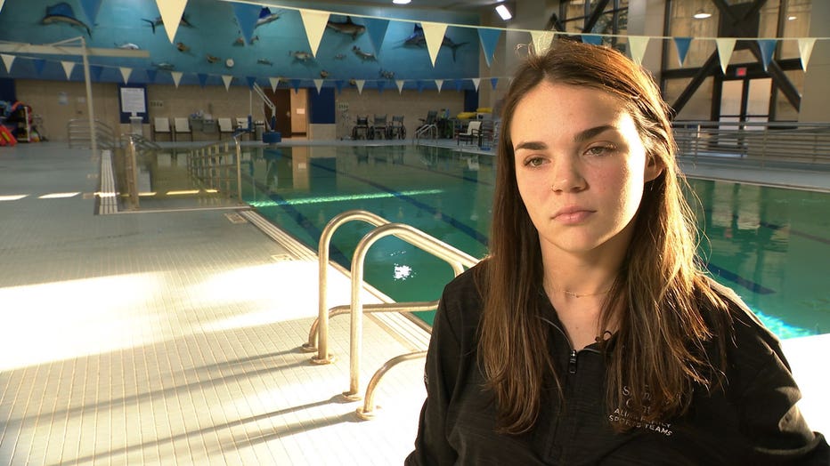 A young woman with long brown hair sits by an indoor swimming pool where she trains.