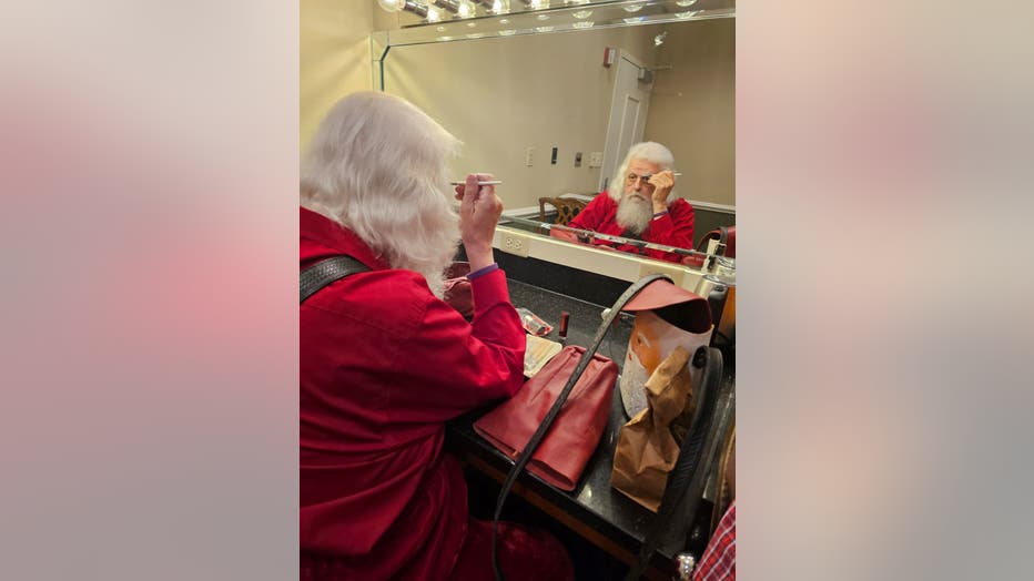 A man playing Santa Claus sits in front of a mirrorv and combs his eyebrows before making an appearance as Santa.