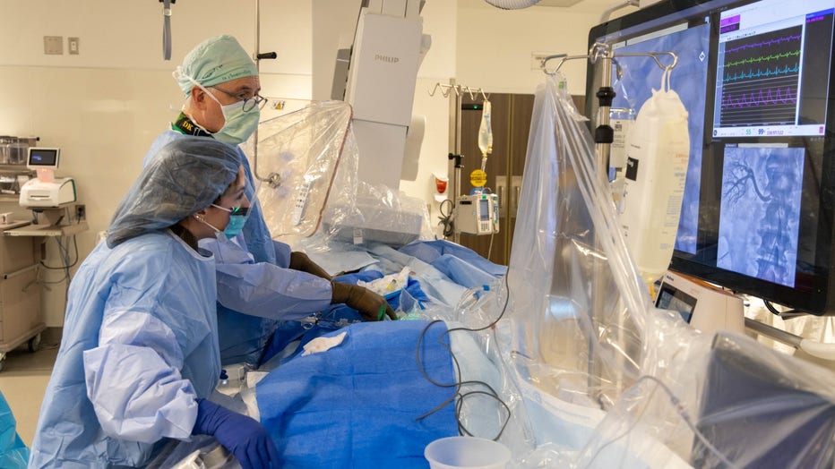 A doctor wearing scrubs and an assistant perform a catheterization procedure