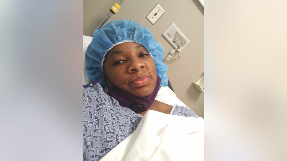 A Black woman takes a selfie of herself before undergoing a surgical procedure. She is wearing a surgical gown and cap and is lying on a gurney.