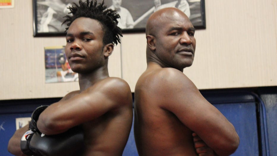 Evan and Evander Holyfield, both in boxing shorts, pose back to back, with their arms crossed.