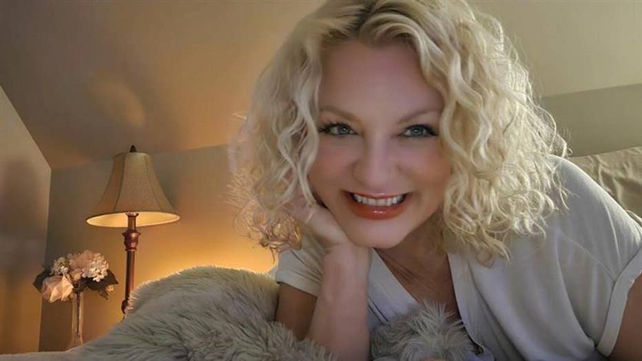 Woman poses for a selfie, smiling and sitting on her bed.