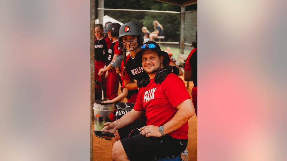 A young softball player poses with her father, her hands on his shoulders