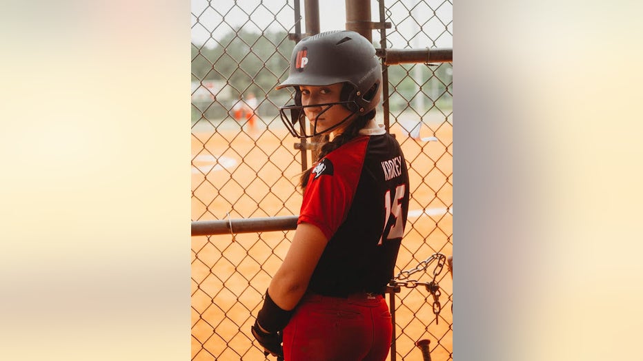 A girl wearing a softball helmet and uniform stands at a fence looking back at the camera. She has a determined expression on her face.