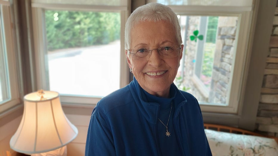 A great haired woman with a short pixie haircut stands on her sunporch and smiles.