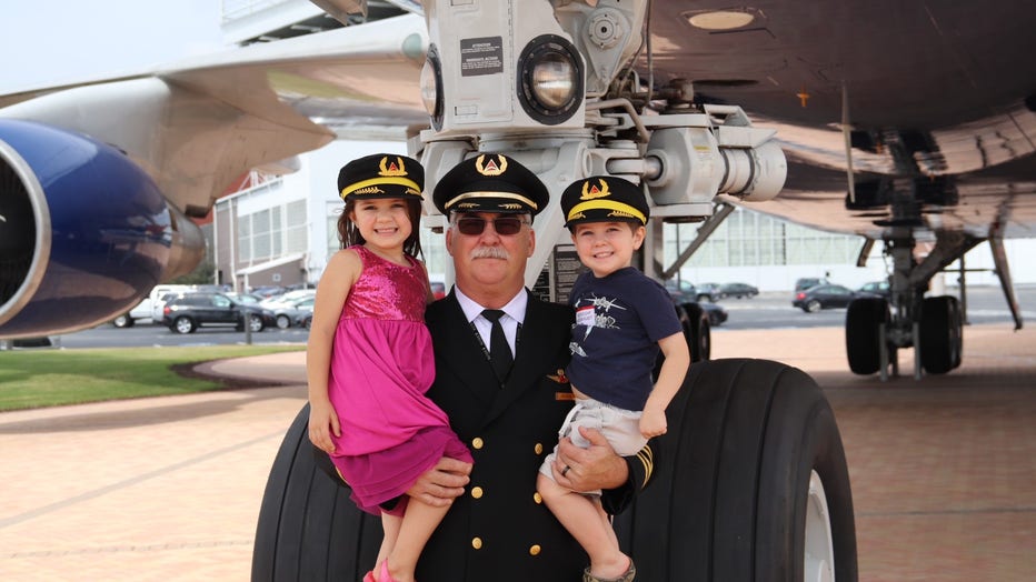 A Delta pilot stands in front of one of the engines of his jet holding two of his young grandchildren in his arms. They are wearing pilot hats.