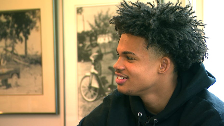 A young man wearing a hoodie smiles as he listens to someone out of the frame