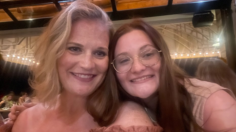 Mother and daughter pose for a photo with their heads leaning in towards one another. Theyre smiling.