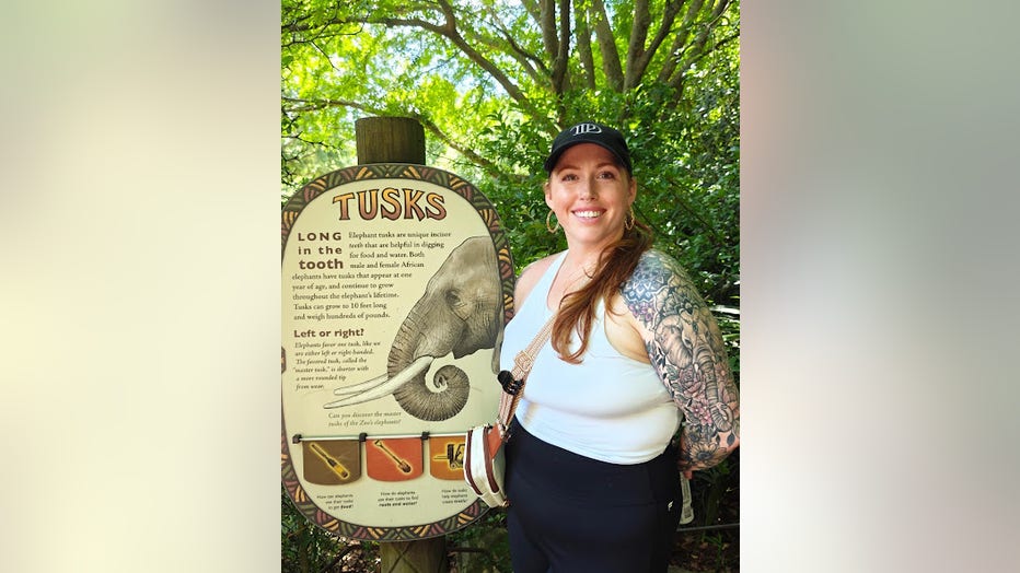 A woman in a baseball hat and tank top stands beside a sign at an elephant enclosure. She's smiling at the camera.