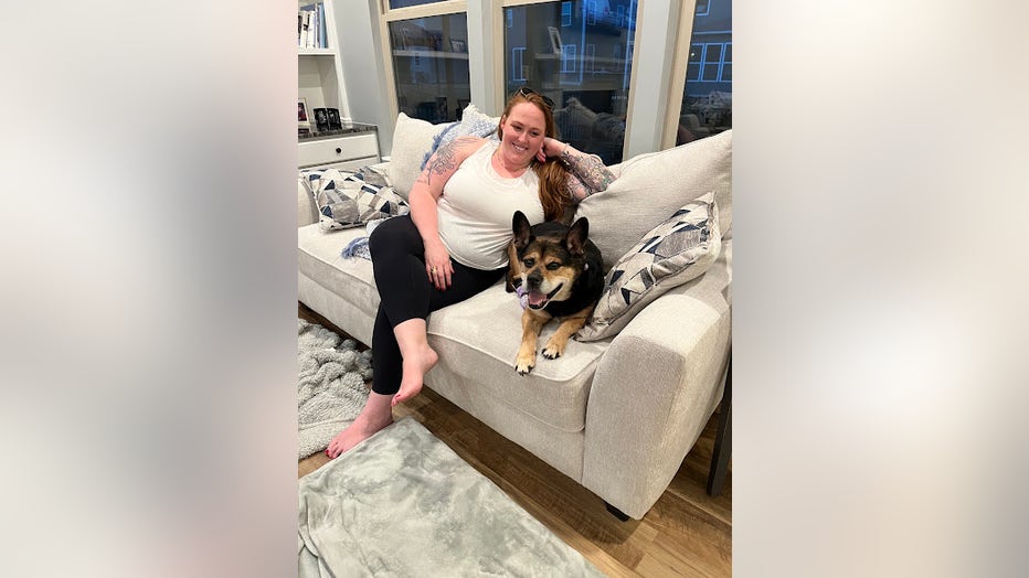 A woman in workout clothes sits on her couch with her dog beside her. They are both looking at the camera and smiling.