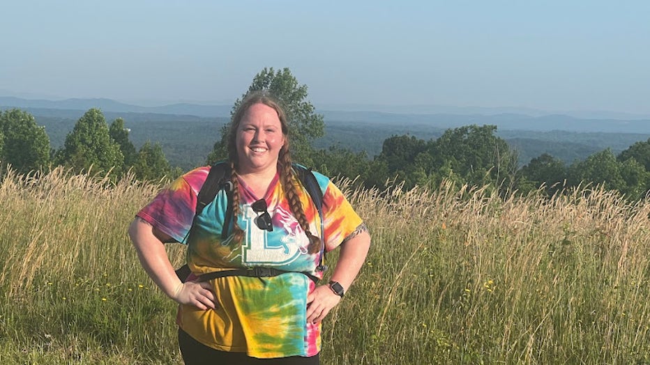 A woman stands with her hands on her hips along a walking trail. She is wearing a backpack and has her hair pulled back. She's smiling and looks confident.