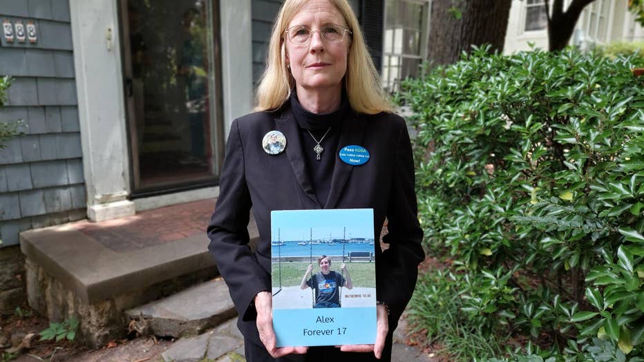 A woman wearing a black suit and glasses stands in front of her home holding a photo of her 17-year-old son, who died by suicide.