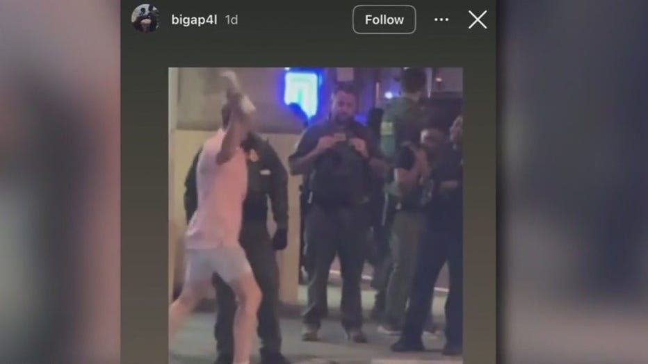 A screenshot of an Instagram story showing a man in a pink t-shirt and white shorts winding up to throw a wrapped deli sandwich in his hand at a man in a law enforcement uniform.
