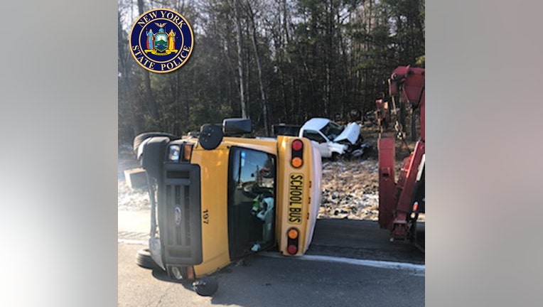 A yellow school bus on its side on the road and a damaged white pickup truck in the background