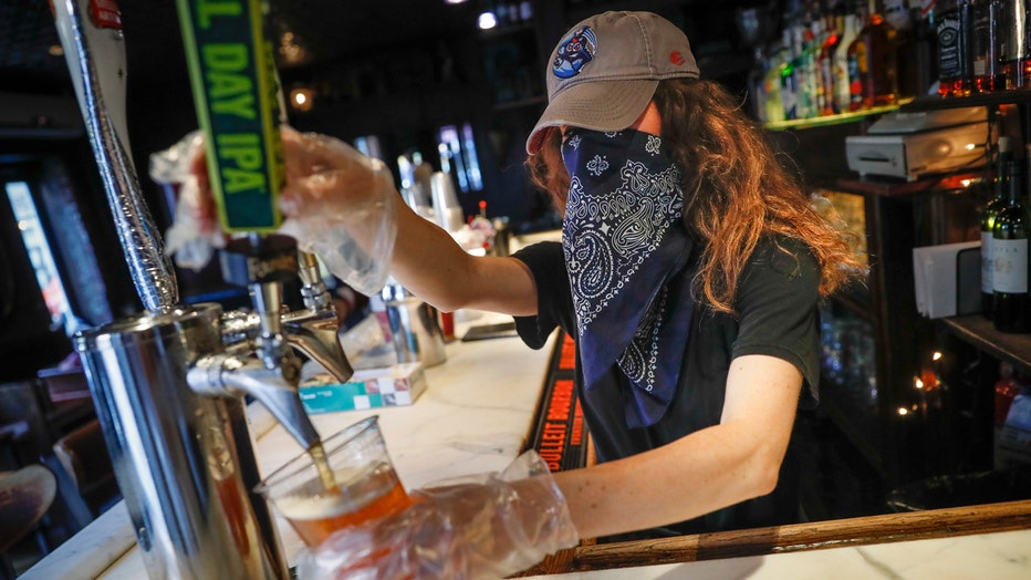 Bartender wearing a baseball cap on her head and a bandanna over her face pours beer into a cup at a bar