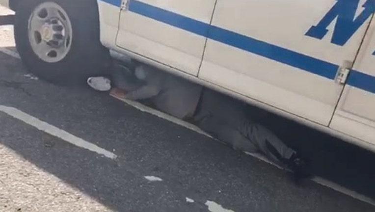 A person wearing a mask lies on the ground under an NYPD vehicle
