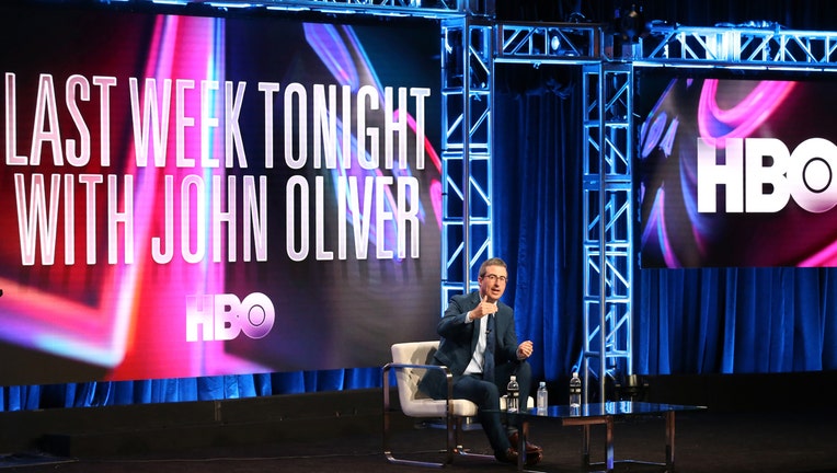 John Oliver sits on stage with large monitors behind him displaying Last Week Tonight with John Oliver and HBO