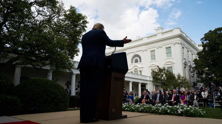 Trump seen from behind stands at a lectern in the Rose Garden as he speaks to several people sitting in chairs