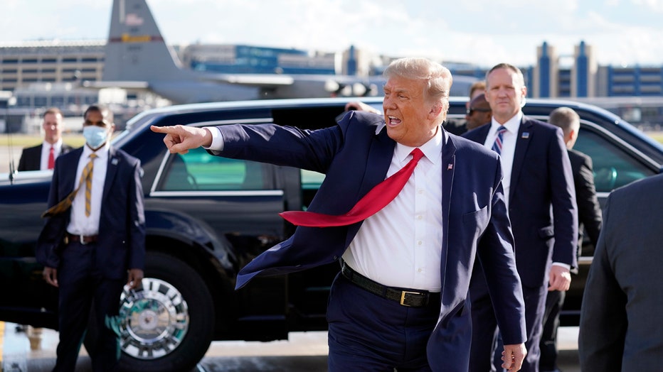 Trump, wearing blue suit and red tie, points to his right as he walks on tarmac; Secret Service agents and a limo are seen behind him